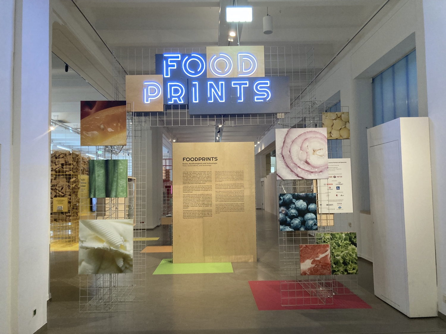 Eingangsbereich zur Ausstellung Foodprints, Schritzug Foodprints in Leuchtbuchstaben über einem aus Gitterboxen zusammengestellten Tor, das mit Detailaufnahmen von Nahrungsmitteln dekoriert ist