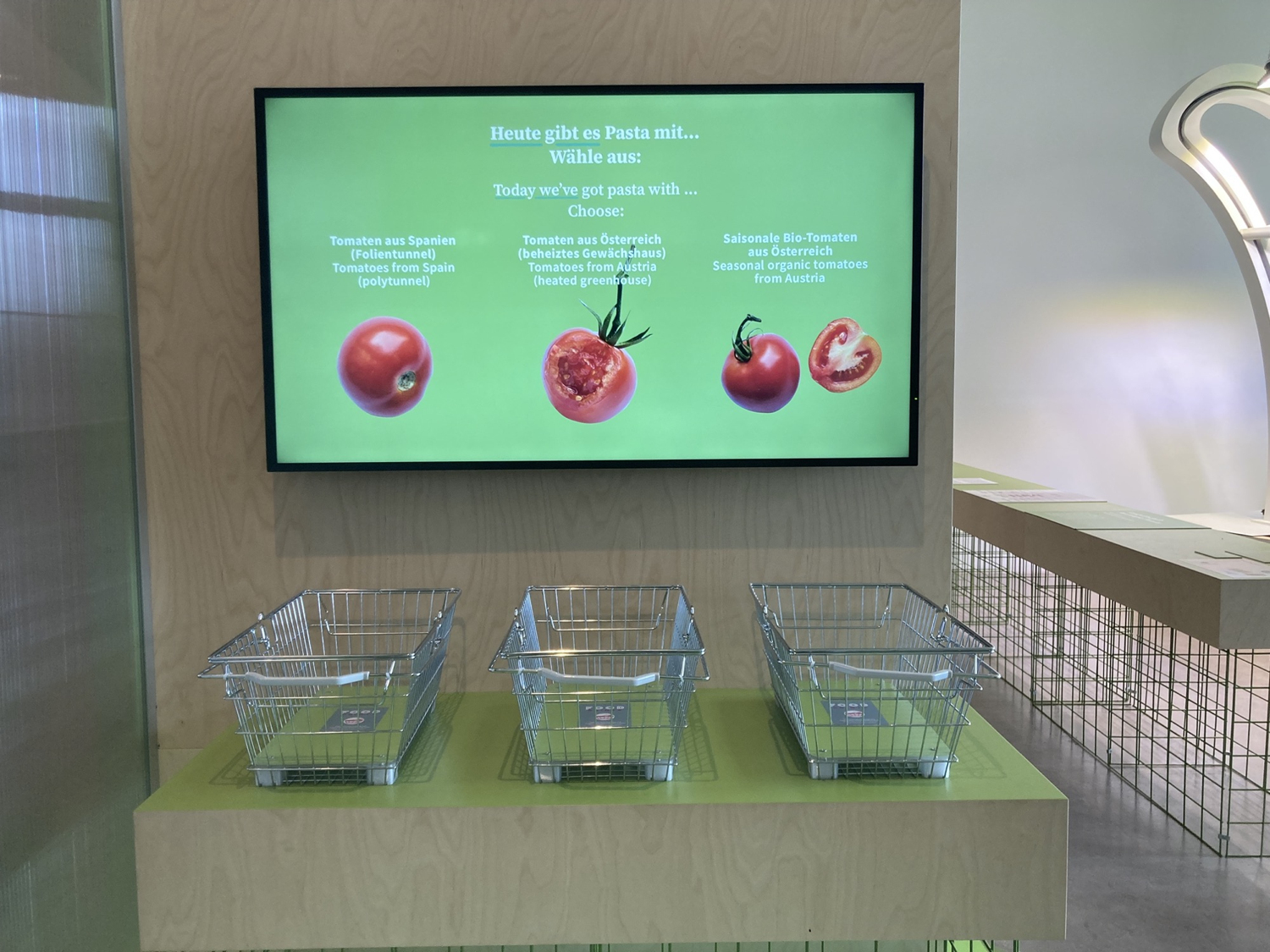 ein Bildschirm, auf dem drei Sorten von Paradeisern/Tomaten abgebildet sind, darunter drei Einkaufskörbe, in denen Besucher:innen ihre Karten ablegen können, um eine Einkaufsentscheidung zu treffen