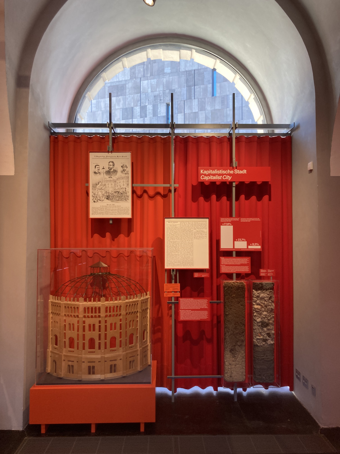Ausstellungsdisplay in einer Nische mit halbkreisförmigem Fenster darüber, vor einem roten Vorhang ist ein Architekturmodell eines Wiener Gasometers und daneben ein Vergleich von Bodenprofilen zu sehen