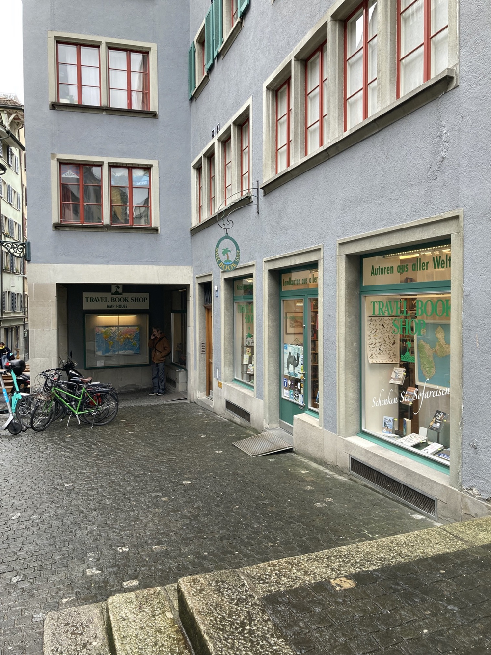 Fassade des Travel Book Shop in Zürich, über dem Eingang hängt ein Zunftzeichen mit einer Palme, die Fensterrahmen sind grün gestrichen, im Hintergrund ist eine Reliefkarte in einem Schaukasten zu sehen