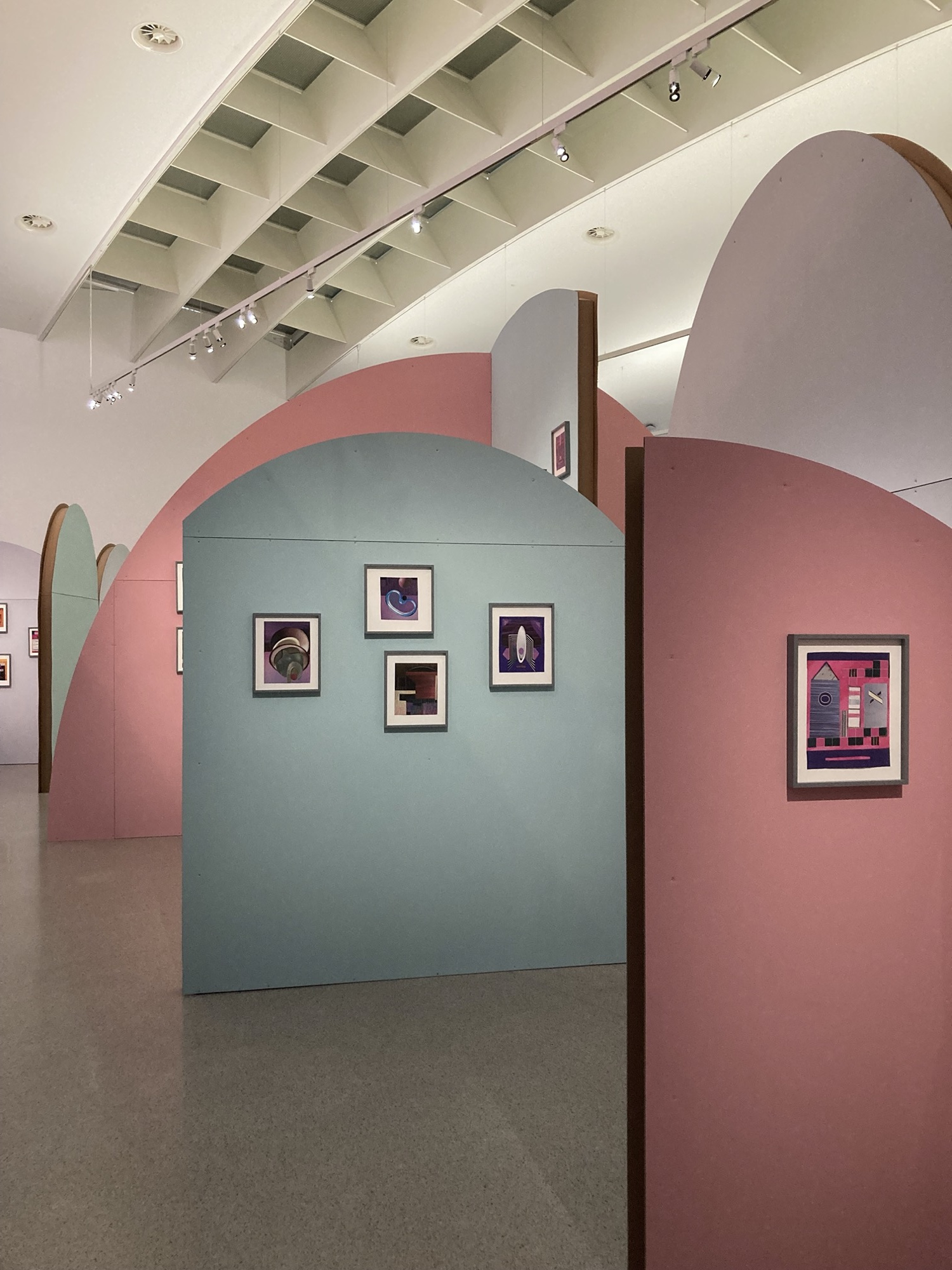 Ausstellungsräumlichkeiten im Mumok, in Pastellfarben gestrichene Wände mit runden Abschlüssen bilden Räume, an den Wänden sind jeweils einige Collagen aufgehängt, rechts vorne ist eine Collage mit pinken und violetten Elementen zu sehen, die ein geometrisches System zeigt mit einem Element, das möglicherweise eine Rakete darstellt