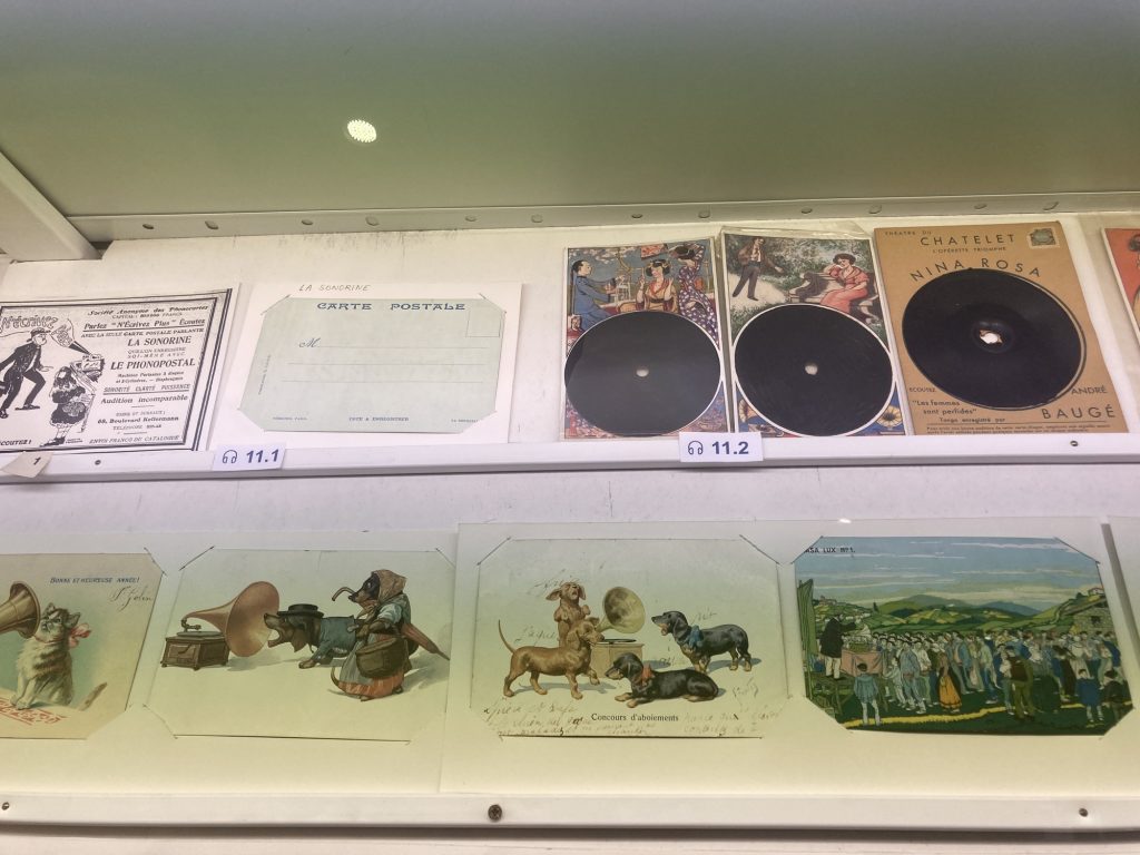 2 Reihen von Postkarten, oben links die Vorder- und Rückseite einer Postkarte, auf der Vorderseite ein französischer Text und eine Schwarz-Weiß-Illustration eines Phonographen, aus dem Sprache erklingt, die Rückseite enthält den Schriftzug „CARTE POSTALE“ und Textfelder zum Eintragen der Adresse, rechts daneben drei Karten, bei denen eine schwarze Scheibe mit einem Loch in der Mitte das zentrale Element bildet, auf der Postkarte ganz rechts ist der Titel „NINA ROSA“, darunter Illustrationen von Katzen und Hunden, die in die Öffnung eines Grammophons hineinhören oder hineinbellen