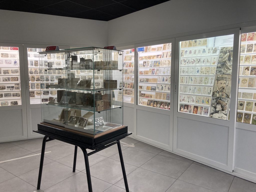 räumliche Ansicht des Postkartenmuseums, im Vordergrund steht ein Tisch mit einem Glaskasten darauf, in dem auf vier Ebenen spezielle Postkartenexponate betrachtet werden sollen, im Hintergrund sind die Wände mit hohen Schaukästen verkleidet, in denen reihenweise verschiedenste Arten von Postkarten ausgestellt sind