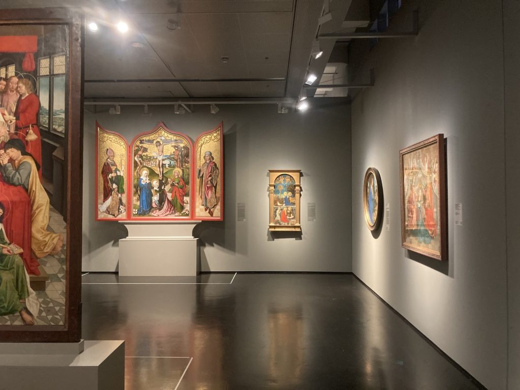 Blick in den Ausstellungsraum, an der hinteren Wand hängt ein dreitiliges Altarbild mit offenen Flügeln, in der Mitte die Kreuzigungsszene, rechts davon ein Bild in einem Säulenrahmen