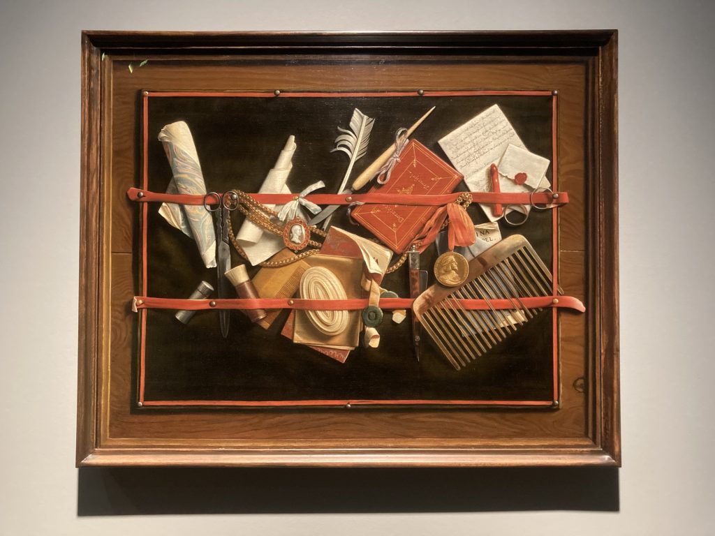 ein brauner Bilderrahmen, das Bild zeigt verschiedene Gegenstände, die von einem roten Band an einer schwarzen Tafel gehalten werden, die Gegenstände (Feder, Schere, Brieföffner, Kette mit Medaillon, rotes Notizbuch, Brief, Kamm) wirken real, obwohl sie nur gemalt sind