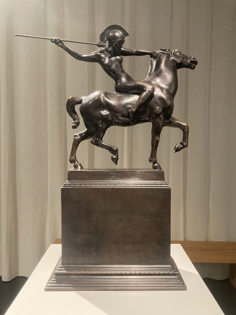 Skulptur aus Bronze, auf einem Pferd reitet eine nackte weibliche Gestalt, sie trägt einen Kriegshelm und hat die rechte Hand mit dem Speer nach hinten gestreckt, mit der linken Hand drückt sie den Kopf des Pferdes zur Seite, damit der Speer freie Bahn haben wird