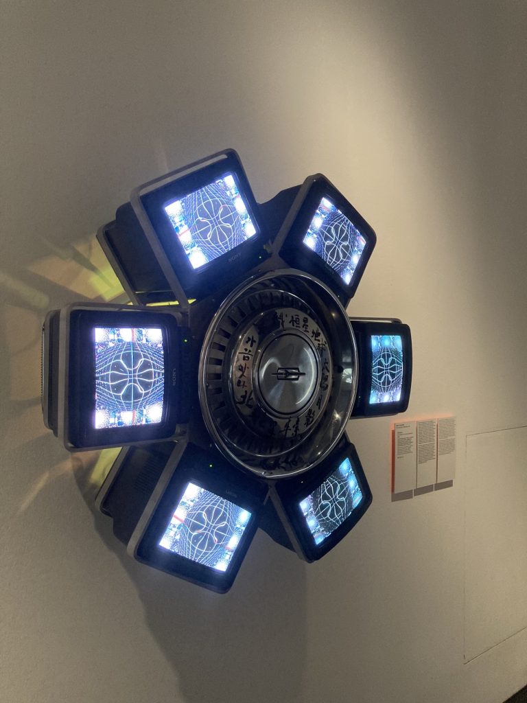 Skulptur aus sechs Monitoren, die kreisförmig um ein Zentrum angeordnet sind, die Mitte hat Ähnlichkeit mit einem Autoreifen ohne den Gummi, sie besteht aus Metall und ist mit koreanischen Schriftzeichen verziert, auf den Bildschirmen ist eine abstrakte Darstellung von Linien zu sehen