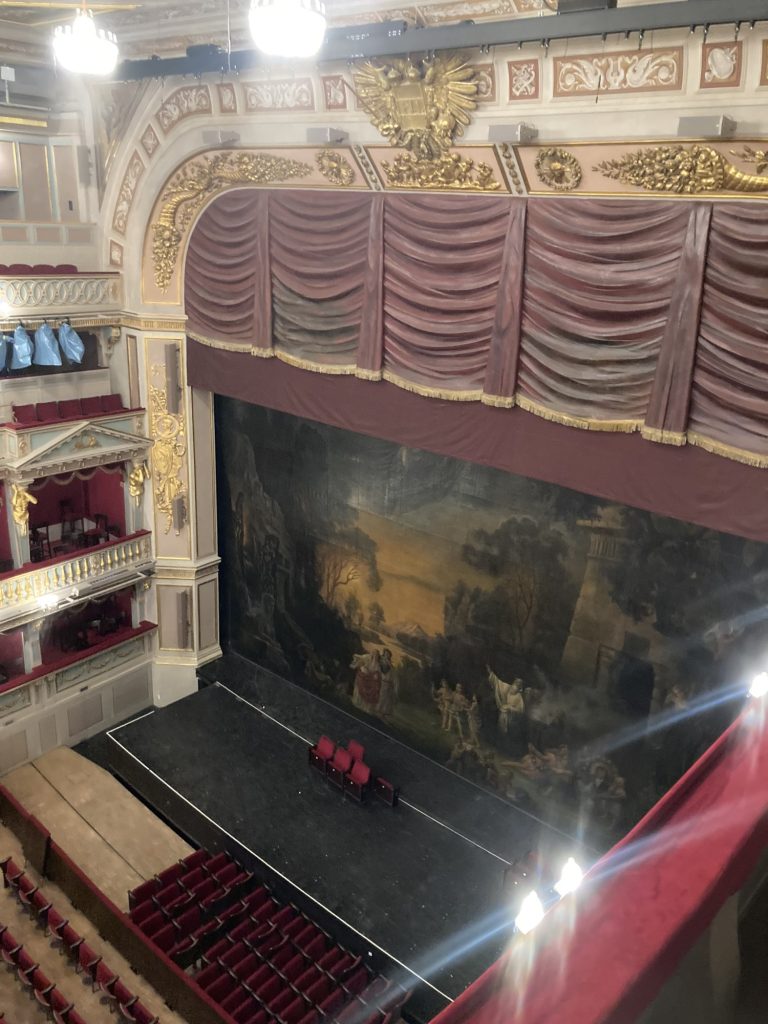 Blick vom Stehplatzbereich im 3. Rang auf die Bühne, der Vorhang ist nun geschlossen, er zeigt eine Theaterszene, auf der Bühne stehen 4 ausgebaute Sitze auf dem mit weißen Strichen markierten Bereich, der das Podest des Orchestergrabens begrenzt
