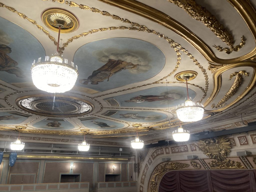 Detailaufnahme der Decke des Zuschauerraums, rund um einen zentralen Lüftungsschacht sind in Form von Blütenblättern Theaterszenen auf blauem Hintergrund gemalt, die Blütenblätter sind mit goldener Dekoration umrankt und von kleinteiligen Kristallleuchtern erhellt