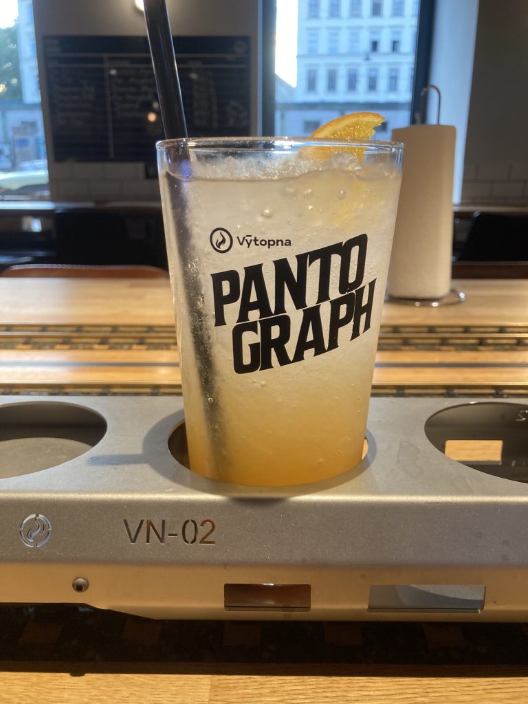 ein großes Glas mit dem Logo Pantograph, das Glas steht in einer Halterung mit zwei freien Plätzen daneben, es enthält unten gelbe Flüssigkeit, die nach oben hin heller wird, einen schwarzen Strohhalm und eine Orangenscheibe, die oben über den Rand schaut
