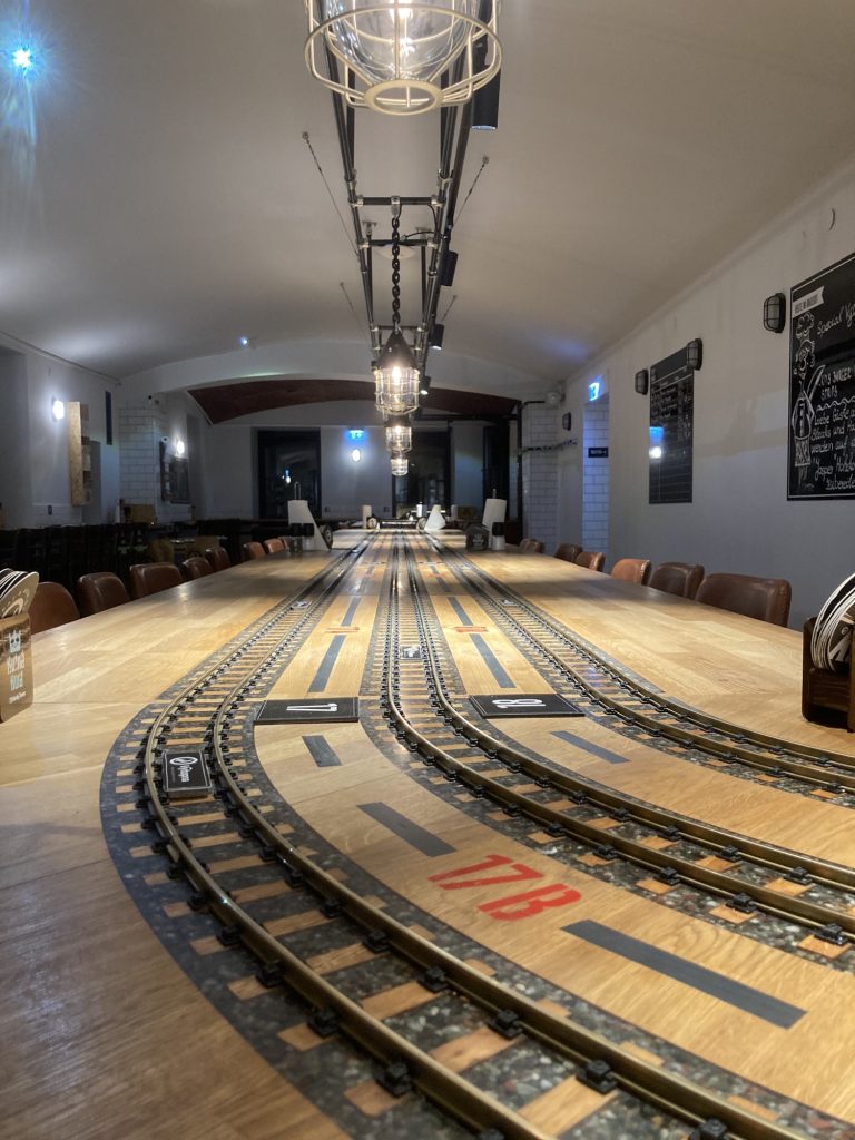 drei Modellbahnschienen nebeneinander auf einem Tisch verlegt, von oben beleuchtet mit in Metallgefäße eingefassten Lampen im Vordergrund verlaufen die Schienen in einer Kurve ins Bild und verschwinden nach hinten ins Bild im Hintergrund, ganz vorne in roter Schrift zwischen den Schienen steht „17B“