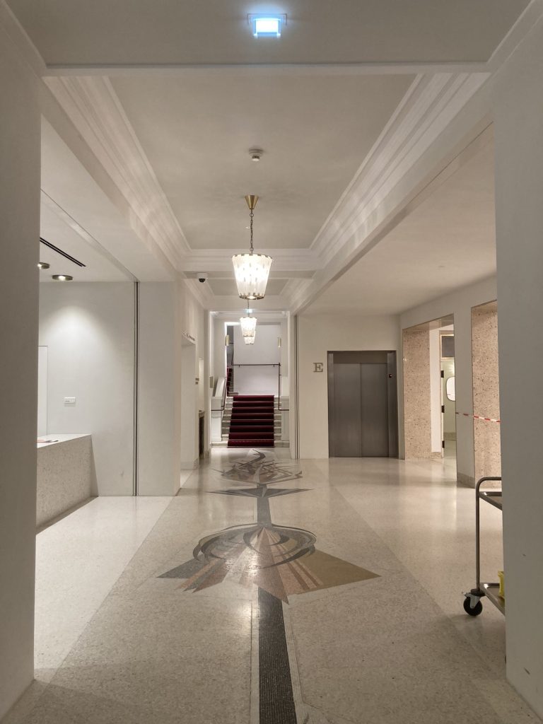 Blick ins renovierte Foyer, den Boden ziert ein aus den 1960er-Jahren stammendes dekoratives Mosaik in Braun- und Grau-Tönen, Wände und Decke erscheinen in weiß und hellen-Grau-Tönen, im Hintergrund führt eine Treppe mit einem roten Teppich nach oben, rechts daneben eine Aufzugstür