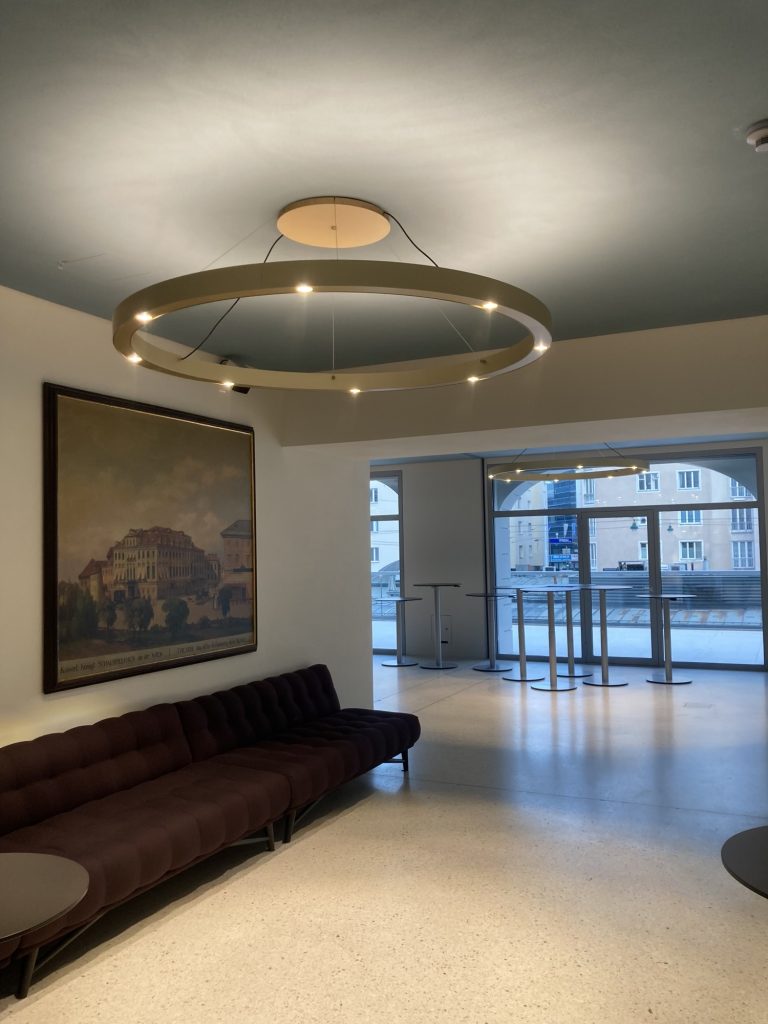 das neue Foyer im ersten Stock ist eine Mischung aus alt und neu, links ein Gemälde, das das Theater in seiner Entstehungszeit zeigt (damals stand es nicht inmitten der Stadt und war nicht von Gebäuden umringt), darüber eine moderne runde Deckenbeleuchtung, ein großer Ring, der gleichzeitig nach oben sowie nach unten beleuchtet, hinten im Bild die Aussicht auf die Terrasse und die Dächer des Naschmarkts
