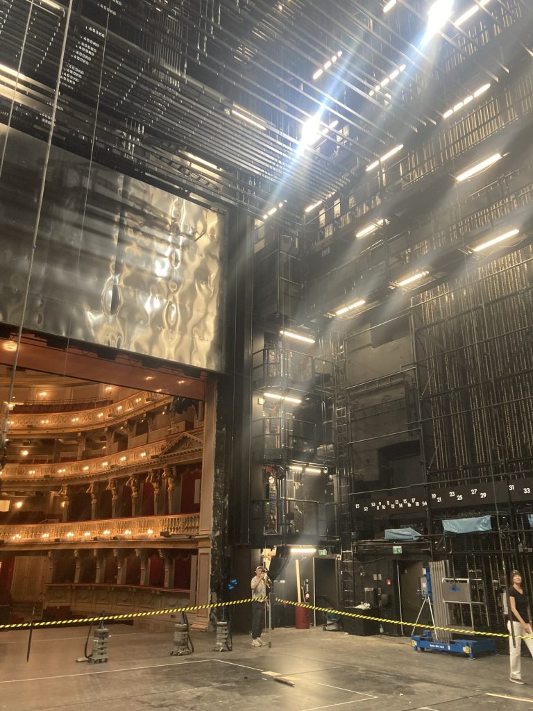 der Bühnenraum des Theaters, links unten ein Ausschnitt des Zuschauerraums, darüber der hochgezogene eiserne Vorhang, rechts der Blick in den Schnürboden, die Scheinwerfer von oben ziehen Lichtschlieren durch das Bild, ein gelb-schwarzes Flatterband grenzt den mittleren Bühnenbereich ab, an einer Ecke dieser Absperrung steht eine Person und fotografiert nach oben