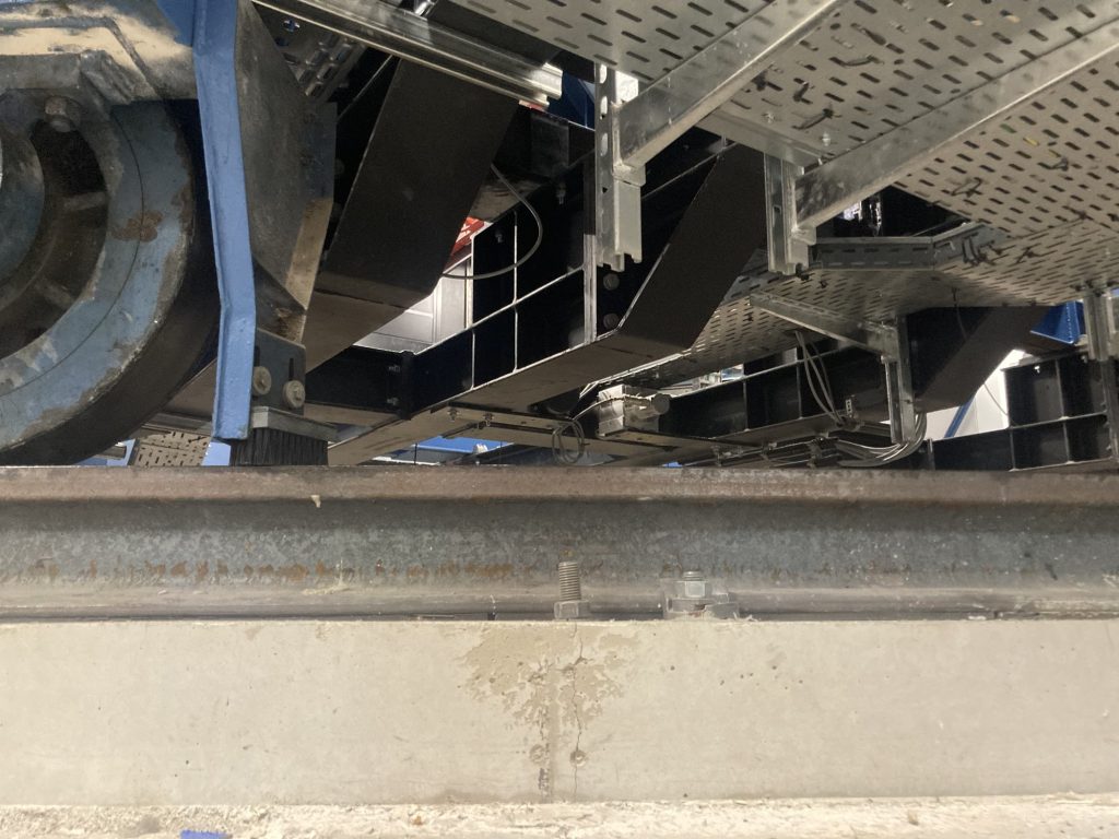 das Fundament der Drehbühne, Eisenbahnschienen sind mit großen Schrauben auf einem gegossenen Betonring verschraubt, links ist ein halbes blaues Rad zu sehen, direkt dahinter wird ein Besen auf den Schienen nachgeführt, über dem Radbereich Kabelschächte 