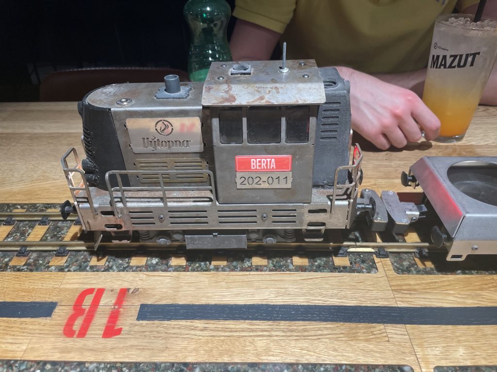 graue Modellbahnlokomotive auf Schienen, am Führhäuschen sind zwei Schilder befestigt, einmal der Name Berta, darunter die Ziffern 202-011, die Lok fährt nach links aus dem Bild, im Hintergrund ist die Hand einer Person neben einem Limonadenglas zu sehen