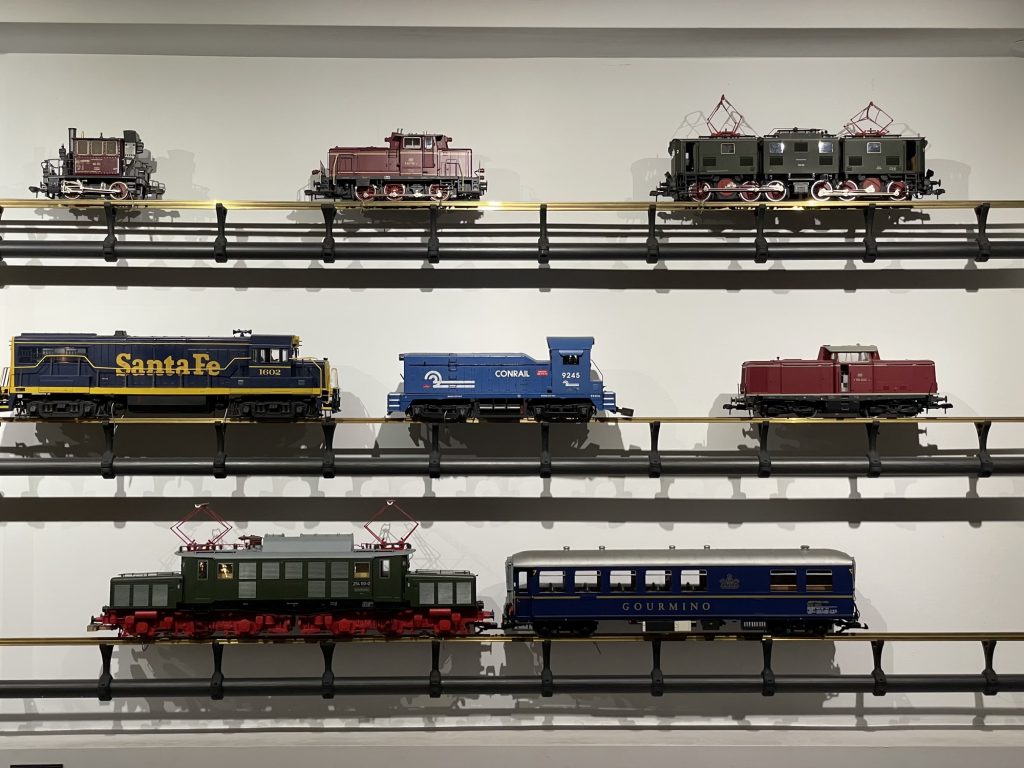 Frontalansicht von 8 Modellbahnlokomotiven bzw. -waggons, die auf an der Wand montierten Schienen ausgestellt sind, 1. Reihe: eine ganz kleine Lok mit 2 Drehgestellen und einem winzigen brauenen Häuschen darauf mit 3 Fenstern, eine etwas größere braune Lok mit 3 Drehgestellen, ein Waggon mit 6 Drehgestellen und 2 Stromabnehmern, 2. Reihe: große dunkelblaue Lok mit der gelben Aufschrift „SantaFe“, kleine hellblaue Lok mit der weißen Aufschrift „Conrail“, rotbraune Lok mit einem winzigen zentralen Führhäuschen, 3. Reihe: große grünliche Lok mit auffälligem rotem Fahrgestell und zwei Stromabnehmern, dahinter ein blauer Passagierwaggon mit der grauen Aufschrit „Gourmino“