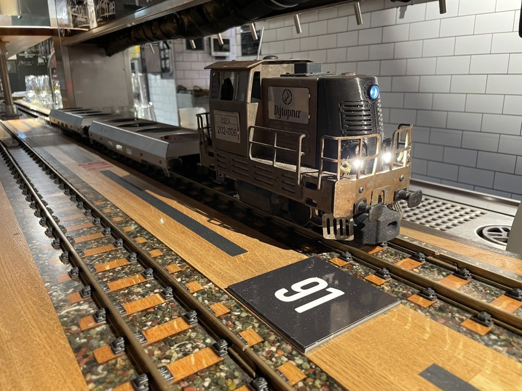 metallisch bronzefarbene Modellbahnlokomotive auf Schienen, sie steht an der Servierstation und zieht zwei Waggons mit jeweils 4 leeren Getränkehaltern, am Führhäuschen sind zwei Schilder befestigt, einmal der Name Pepa, darunter die Ziffern 202-006, die Lok fährt nach rechts aus dem Bild, nach vorne leuchten drei LEDs als Zugspitzensignal, das oberste hat einen bläulichen Schein