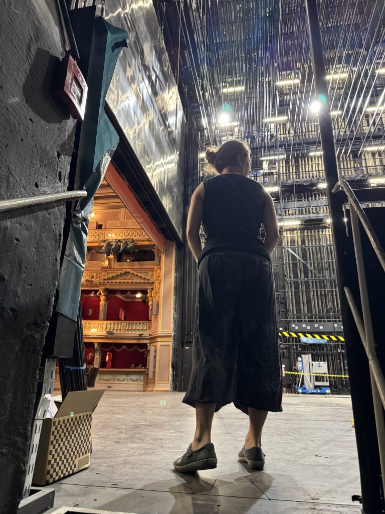 eine Person steht mit dem Rücken zur Kamera am seitlichen Rand einer Bühne, links ist unter dem eisernen Vorhang ein Stück des Zuschauerraums zu sehen, rechts und oben die leeren Kulissenzüge