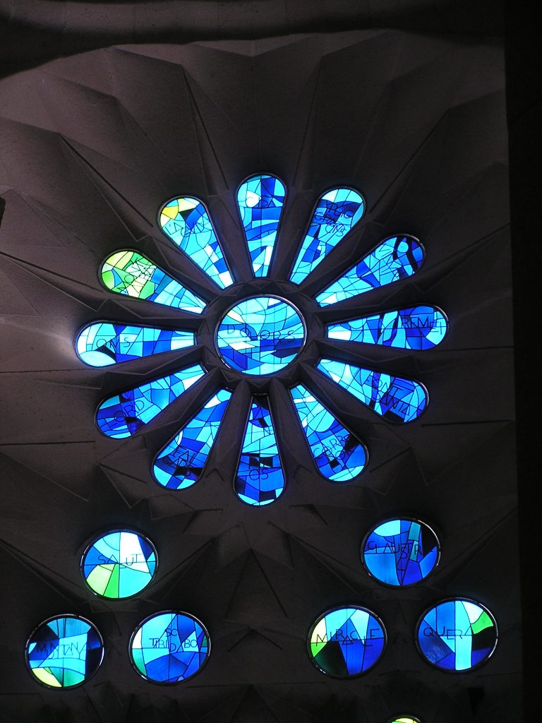 buntes Glasfenster in Blautönen, oben ein Blütenblatt mit 12 Blüten, darunter 6 Kreise jeweils in 3er-Gruppen