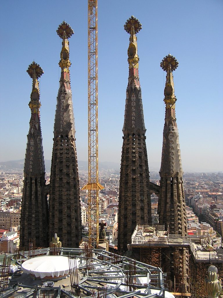 eine Kirchenbaustelle, 4 Türme und dazwischen ein Kran überragen eine Großstadt, im Vordergrund eine runde Plattform mit einem Metallgerüst