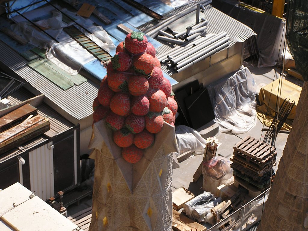 eine Turmspitze mit einer Dekoration aus Erdbeeren, die aus Mosaiksteinen zusammengesetzt sind, dahinter eine Baustelle mit verschiedenem Material