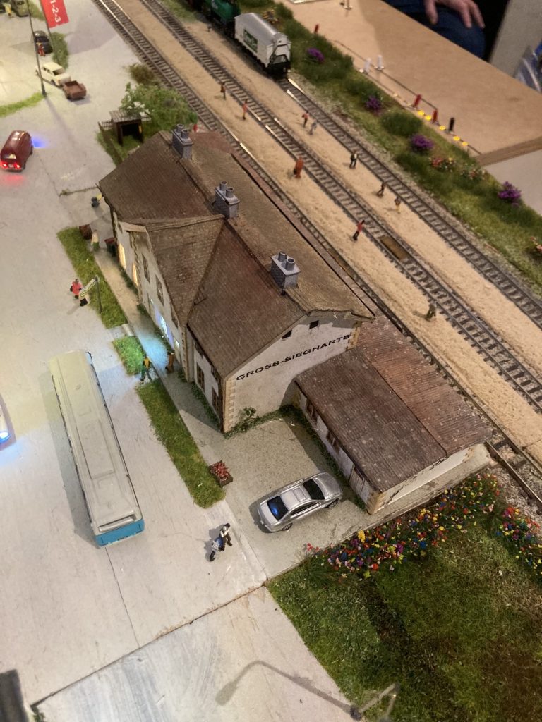 Modell des Bahnhofsgebäudes in Gross-Siegharts, neben dem Gebäude drei Gleise mit Schotterbahnsteigen und verteilten Personen darauf, vor dem Bahnhofsgebäude steht ein Autobus