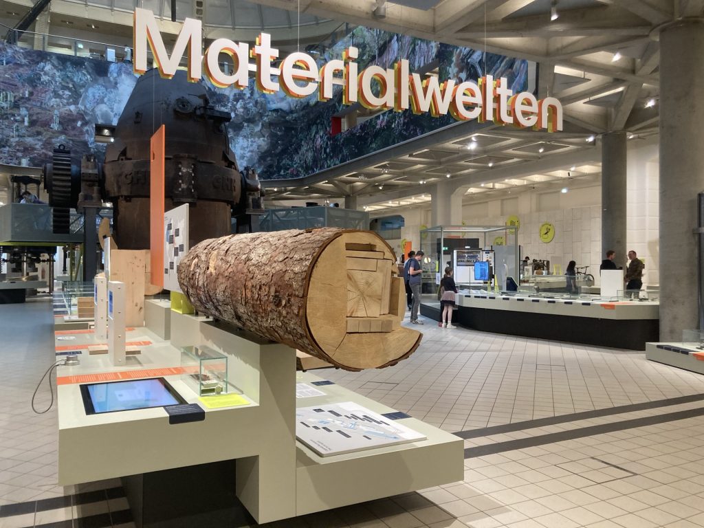 Überblick zur Ausstellung Materialwelten mit dem Titel von der Decke hängend, zentral im Vordergrund ein aufgeschnittener Baumstamm, der die Verarbeitung in einzelne Holzbalken verschiedener Größe veranschaulicht