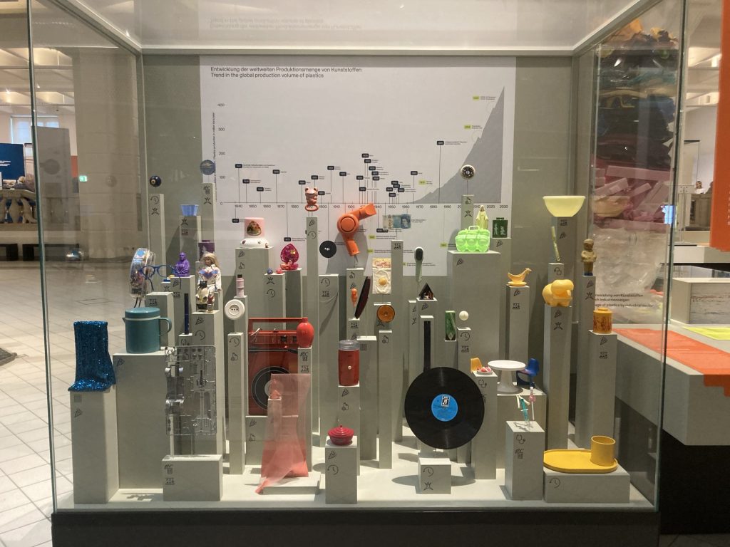 Glaskasten mit Kunststoffprodukten in verschiedenen Farben auf Säulen, diese veranschaulichen den weltweiten Anstieg der Kunststoffproduktion im Zeitverlauf